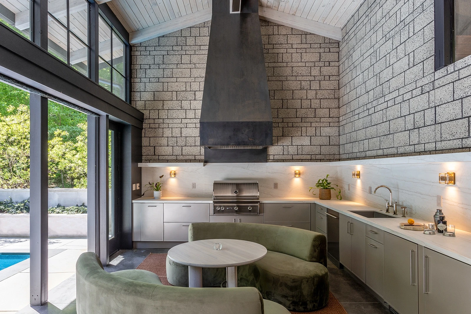Grey outdoor kitchen cabinetry and grill in a glass-walled poolside pavilion