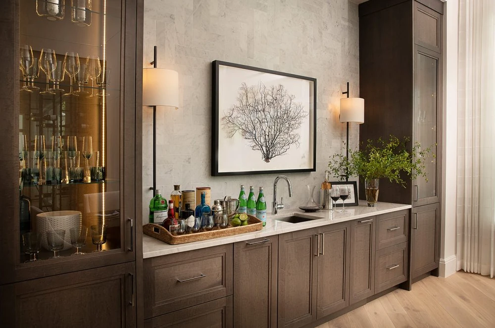 Soft taupe wet bar cabinetry with glass-front storage and a pale stone counter